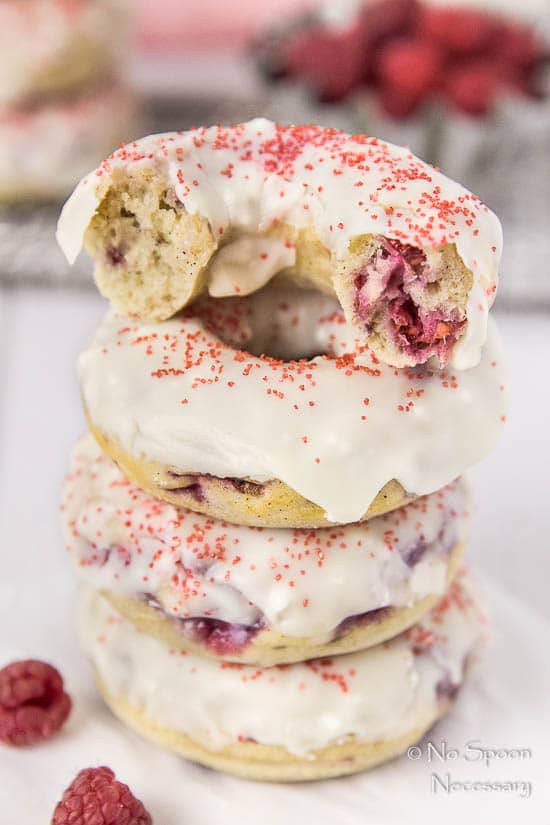 Straight on, up-close shot of a stack of Raspberry Vanilla Baked Donuts with White Chocolate Glaze with the top donut ripped in half; there is a tin of fresh raspberries and additional donuts blurred in the background.