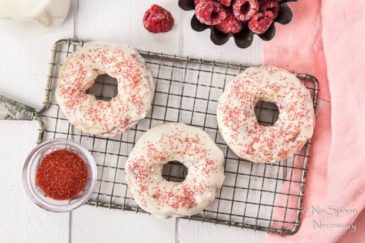 Overhead shot of Raspberry Vanilla Baked Donuts with White Chocolate Glaze on a small wire rack with ramekin of red sprinkles, fresh raspberries, glass jar of milk and a pink linen surrounding the donuts.
