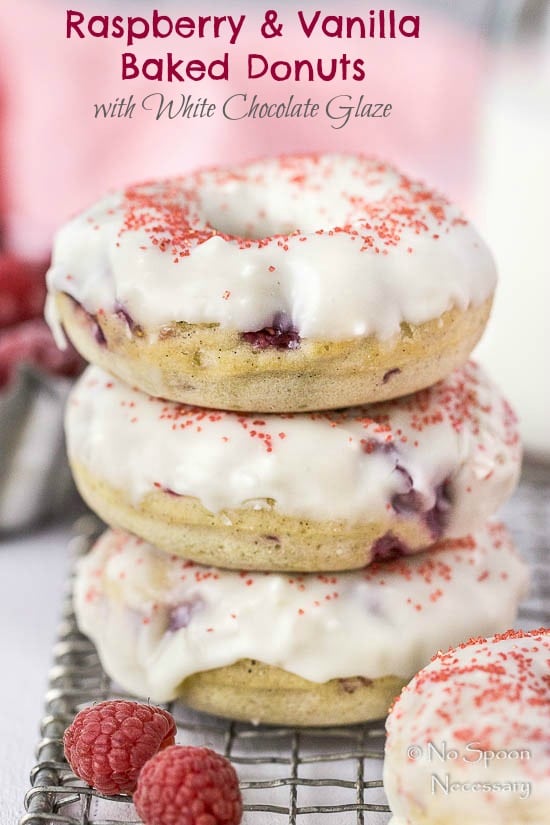 Straight on shot of a stack of Raspberry Vanilla Baked Donuts with White Chocolate Glaze on a small wire rack with a couple fresh raspberries in front of the stack and a glass jar of milk and pink linen blurred in the background.