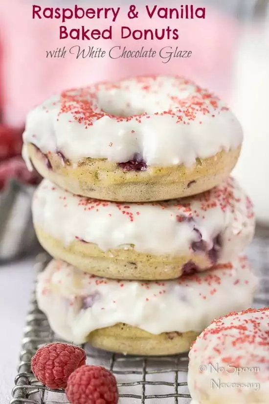 Straight on shot of a stack of Raspberry Vanilla Baked Donuts with White Chocolate Glaze on a small wire rack with a couple fresh raspberries in front of the stack and a glass jar of milk and pink linen blurred in the background.