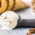 Straight on shot of No Churn Salted Caramel Ripple Butter Pecan Ice Cream in an ice cream scoop laying on a gray surface with waffle cones blurred in the background.
