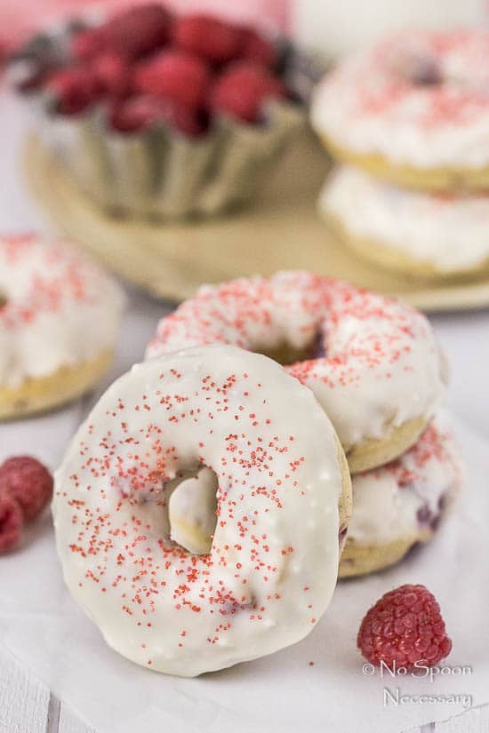 Straight on shot of a stack of two Raspberry Vanilla Baked Donuts with White Chocolate Glaze with a donut leaned up against the stack and a tin of fresh raspberries plus additional donuts blurred in the background. 