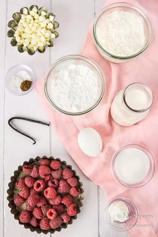 Overhead shot of all the ingredients needed to make Raspberry Vanilla Baked Donuts neatly arranged on a pale purple wood surface with a pink linen.