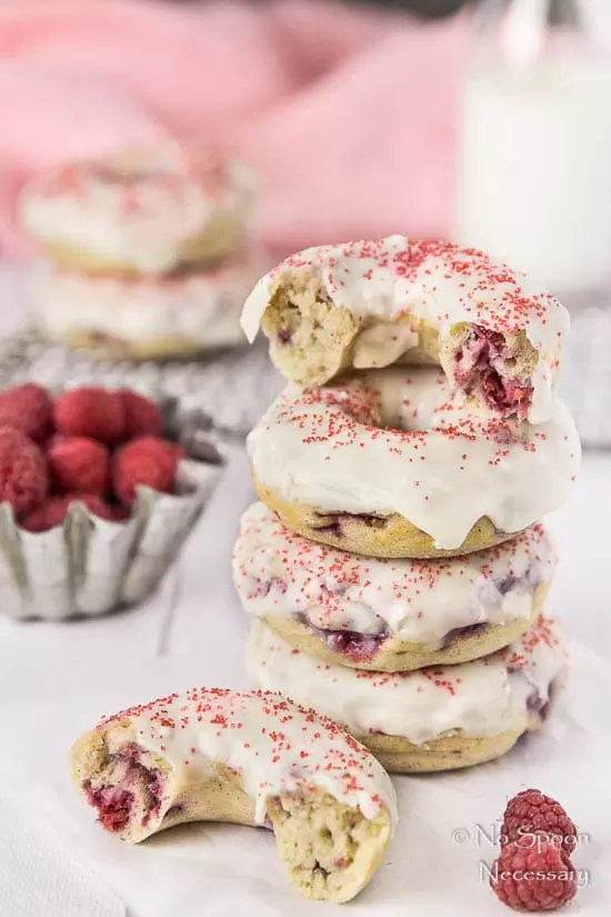 Straight on shot of a stack of Raspberry Vanilla Baked Donuts with White Chocolate Glaze with the top donut ripped in half; there is a tin of fresh raspberries, a glass jar of milk, pink linen and additional donuts blurred in the background.