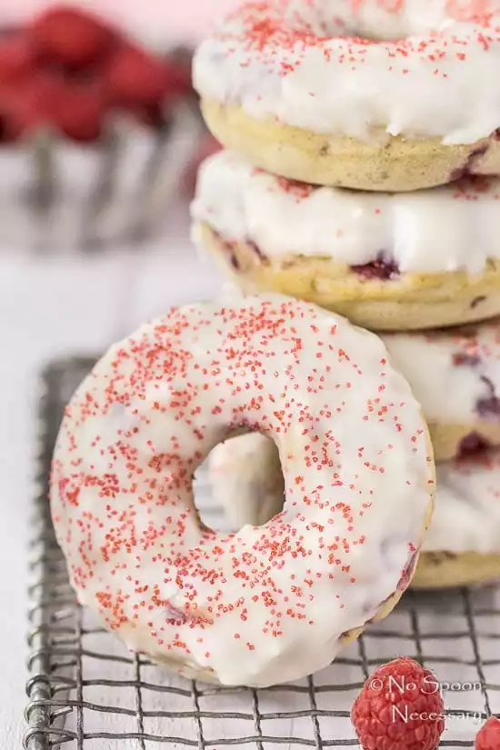 Straight on shot of a stack of Raspberry Vanilla Baked Donuts with White Chocolate Glaze on a small wire rack with one donut leaned up against the stack and a tin of fresh raspberries blurred in the background.