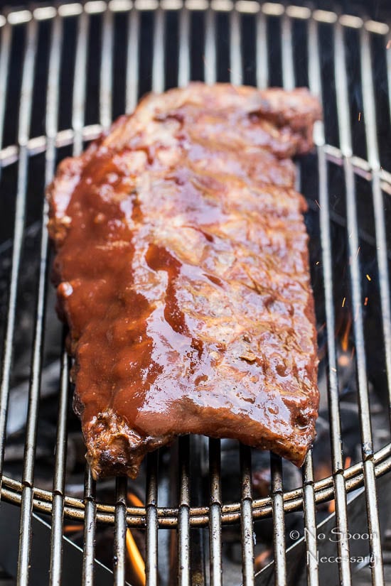 Angled shot of Peach Sriracha BBQ Baby Back Ribs on a charcoal grill - the ribs have been heavily basted with BBQ sauce. (photo of step 7 of the recipe)
