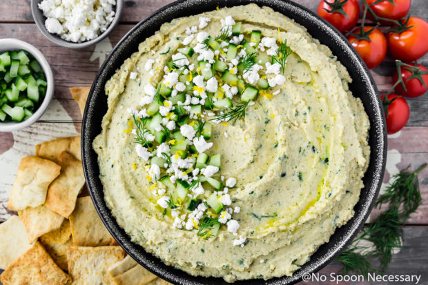 Overhead, landscape shot of a black bowl filled with Easy Tzatziki Hummus Dip with cherry tomatoes, pita chips and bowls of chopped cucumber and crumbled feta surrounding the bowl.