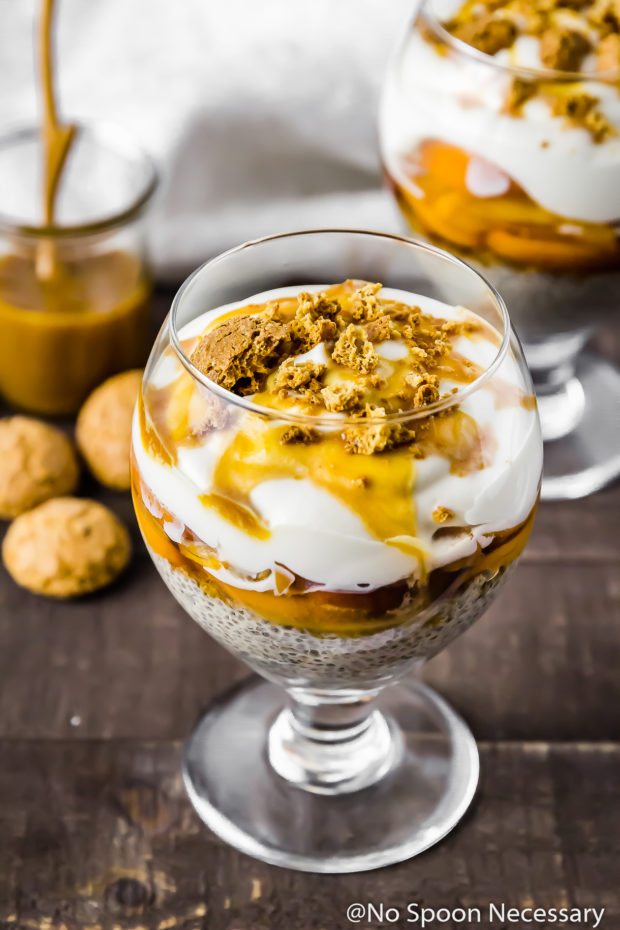Angled shot of a Amaretto Peaches & Cream Chia Pudding Trifle in a stemmed glass with amaretti cookies and an additional trifle blurred in the background.