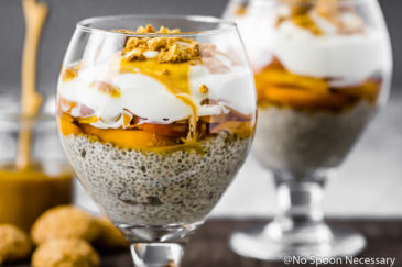 Straight on shot of a Amaretto Peaches & Cream Chia Pudding Trifle in a stemmed glass with amaretti cookies, a jar of caramel and an additional trifle blurred in the background.