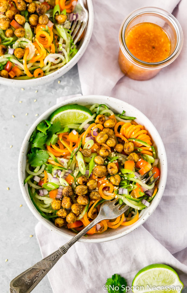 Overhead shot of Thai Roasted Chickpeas with Vegetable Noodle Salad in a small bowl with a fork inserted into the noodles and an additional salad, jar of dressing, pale purple linen and lime wedge surrounding the bowls.