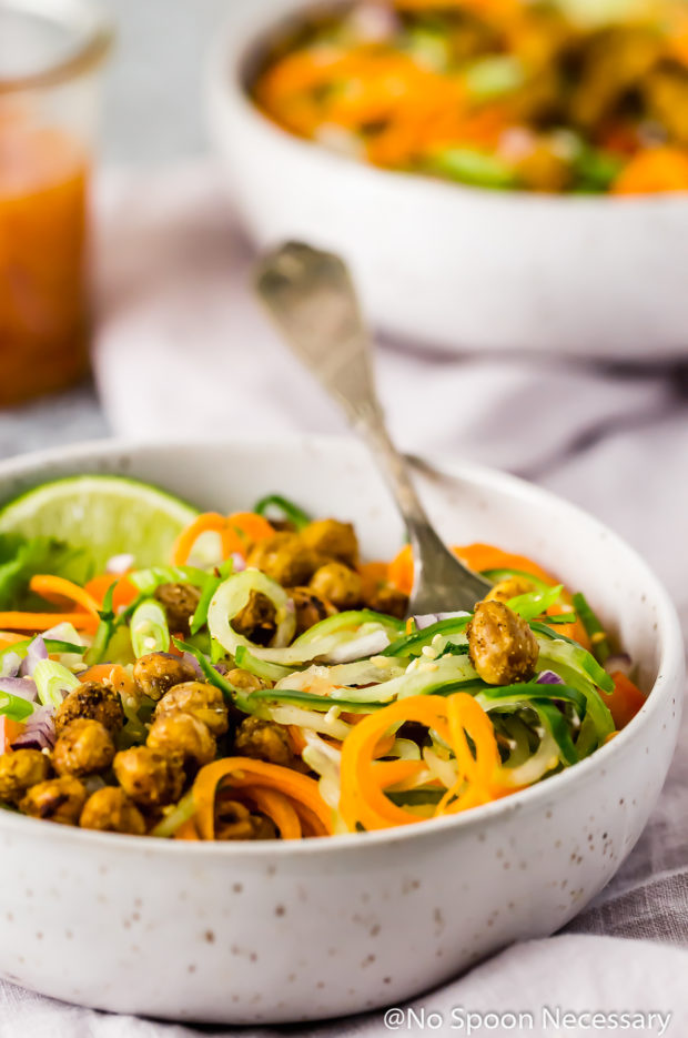 Angled shot of Thai Roasted Chickpeas with Vegetable Noodle Salad in a small bowl with a fork inserted into the noodles and an additional salad and jar of dressing blurred in the background.