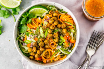 Overhead shot of Thai Roasted Chickpeas with Vegetable Noodle Salad in a small bowl with a jar of dressing, pale purple linen, forks and lime wedge surrounding the bowl.