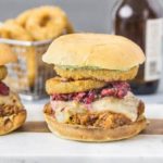 Straight on, landscape shot of three Raspberry BBQ Pulled Pork Sandwich with fresh raspberry salsa and onion rings on wood board with a basket of onion rings and beer bottles blurred in the background.