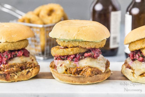 Straight on, landscape shot of three Raspberry BBQ Pulled Pork Sandwich with fresh raspberry salsa and onion rings on wood board with a basket of onion rings and beer bottles blurred in the background.