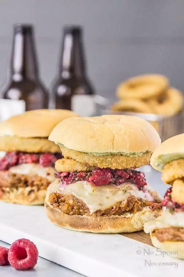 Straight on shot of three Raspberry BBQ Pulled Pork Sandwich with fresh raspberry salsa and onion rings on wood board with a basket of onion rings and beer bottles blurred in the background.