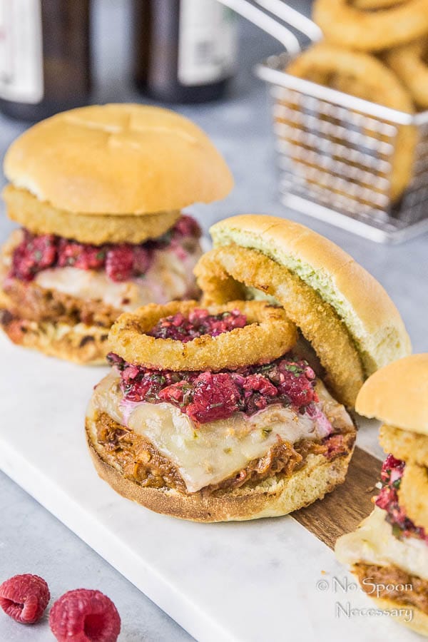 Slightly angled shot of Raspberry BBQ Pulled Pork Sandwiches with fresh raspberry salsa and onion rings on a white serving board with a basket of onion rings blurred in the background.
