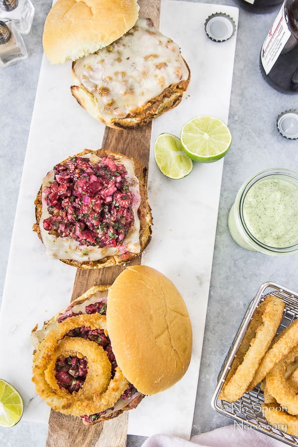 Overhead shot of three Raspberry BBQ Pulled Pork Sandwich with Raspberry Salsa & Cilantro-Lime Aioli on a white serving board with a basket of onion rings, lime wedges and beer bottles surrounding the board.