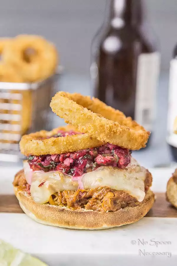 Straight on shot of a Raspberry BBQ Pulled Pork Sandwich with fresh raspberry salsa and onion rings on a white plate with a basket of onion rings and beer bottles blurred in the background.
