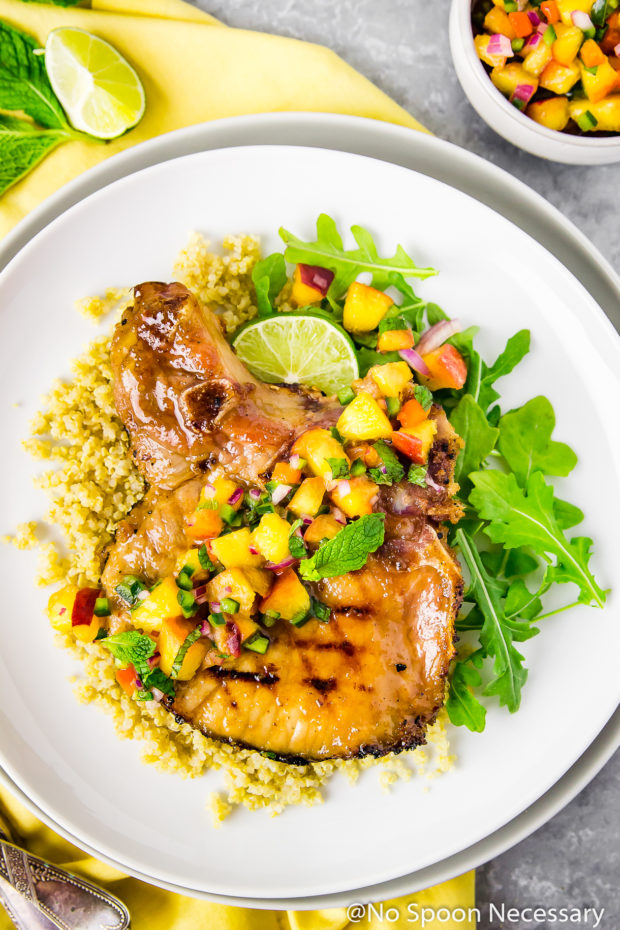 Overhead shot of Ginger Honey Glazed Pork Chops with Peach-Poblano Salsa on quinoa and arugula with a lime wedge on a white plate with a yellow linen under the plate and a bowl of salsa tucked in the corner.