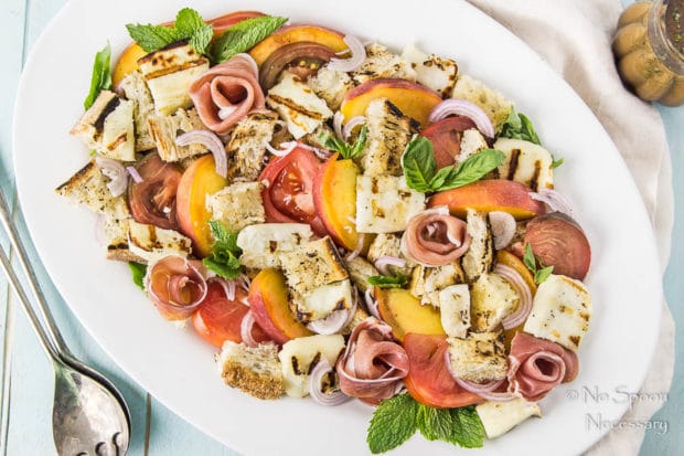 Overhead shot of Grilled Halloumi Peach Panzanella with Prosciutto on a white platter with a jar of basil vinaigrette and serving spoons surrounding the platter.