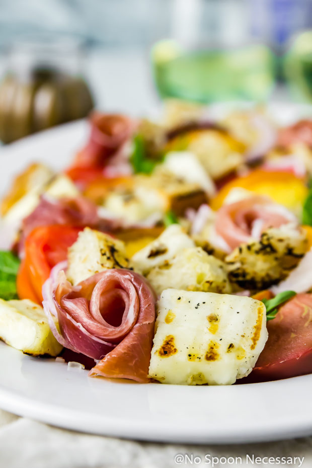 Angled shot of Grilled Halloumi Peach Panzanella with Prosciutto on a white platter with the focus of the shot of a slice of prosciutto and halloumi; with a jar of basil vinaigrette and wine glass blurred in the background.