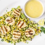 Overhead, landscape shot of Grilled Scallops with Avocado & Corn Salsa on a white platter with a small bowl of Honey-Lime Vinaigrette, fresh cilantro and lime wedges.