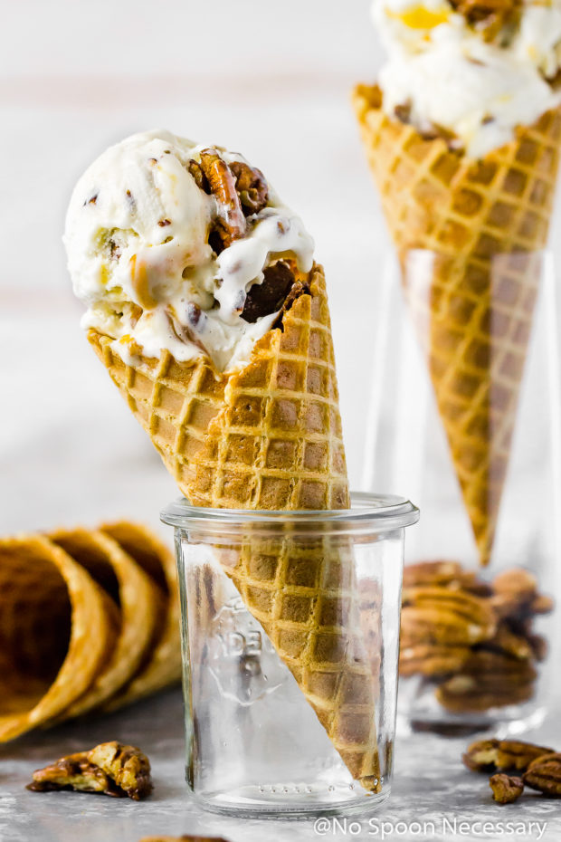 Straight on shot of No Churn Salted Caramel Ripple Butter Pecan Ice Cream in an ice cream cone standing up in a glass jar with an additional cone of ice cream blurred behind it.