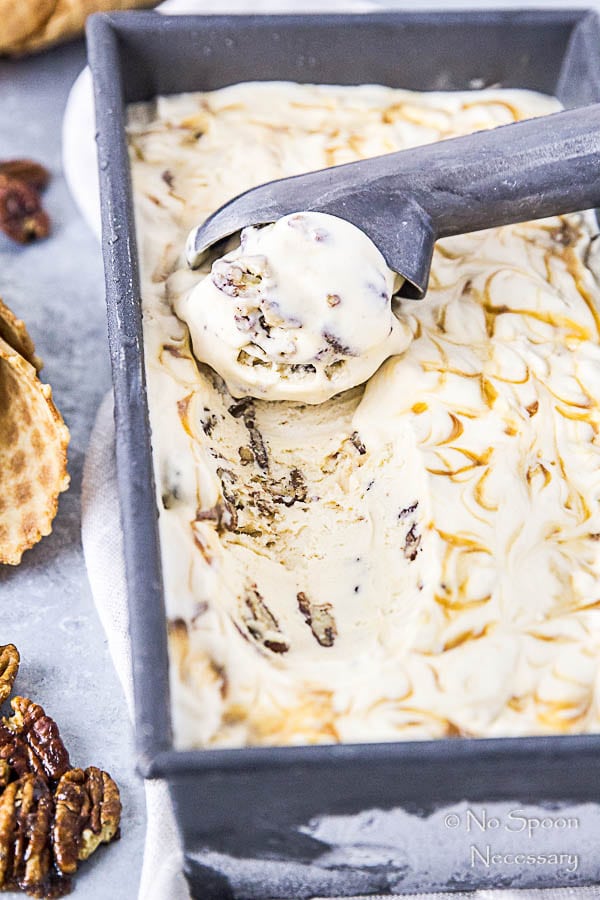 Angled shot of No Churn Salted Caramel Ripple Butter Pecan Ice Cream in a silver container with an ice cream scoop and there are ice cream cones and buttered pecans surrounding the container.