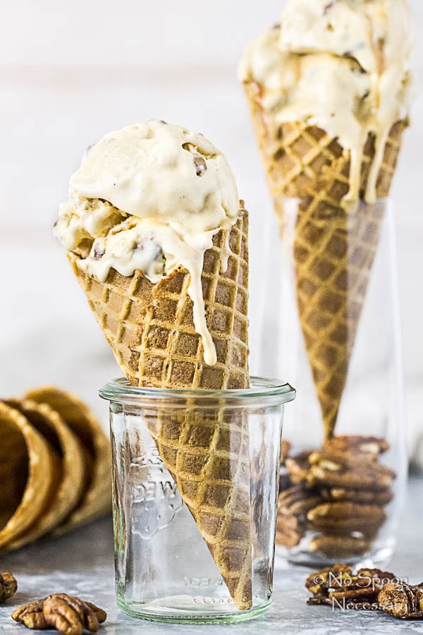 Straight on shot of a tall glass jar holding a No Churn Salted Caramel Ripple Butter Pecan Ice Cream melting in a waffle cone with an additional cone of ice cream blurred behind it.