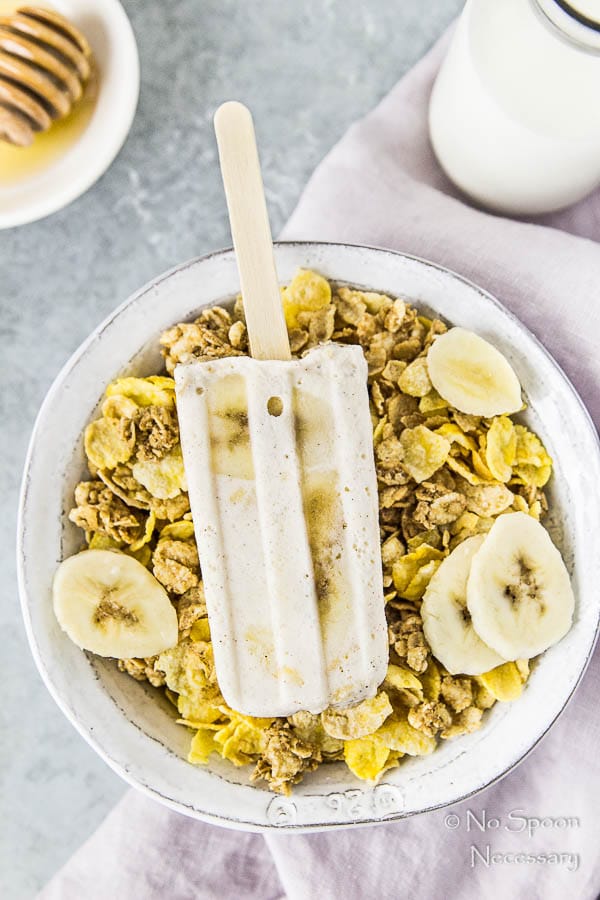 Overhead shot of Banana & Honey Cereal Breakfast Popsicles on top of a bowl of cereal with a purple linen under the bowl and a jar of milk and ramekin of honey in the upper corners of the shot.