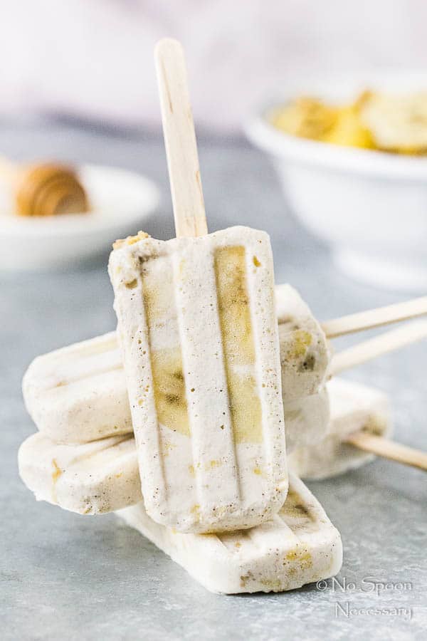 Straight on shot of a stack of Banana & Honey Cereal Breakfast Popsicles on a gray surface with a bowl of cereal and ramekin of honey blurred in the background.