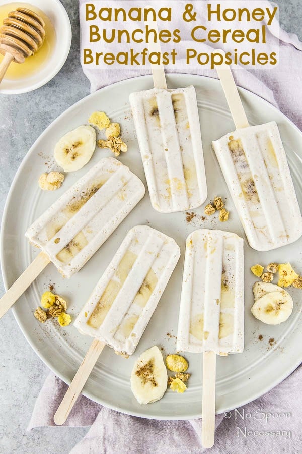 Overhead shot of Banana & Honey Cereal Breakfast Popsicles on a gray plate with slices of bananas and clusters of cereal with a purple linen under the plate and a white ramekin of honey in the corner.