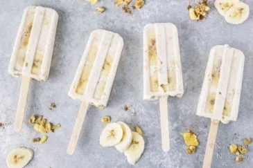 Overhead shot of Banana & Honey Cereal Breakfast Popsicles on a gray surface with slices of bananas and clusters of cereal surrounding the popsicles.