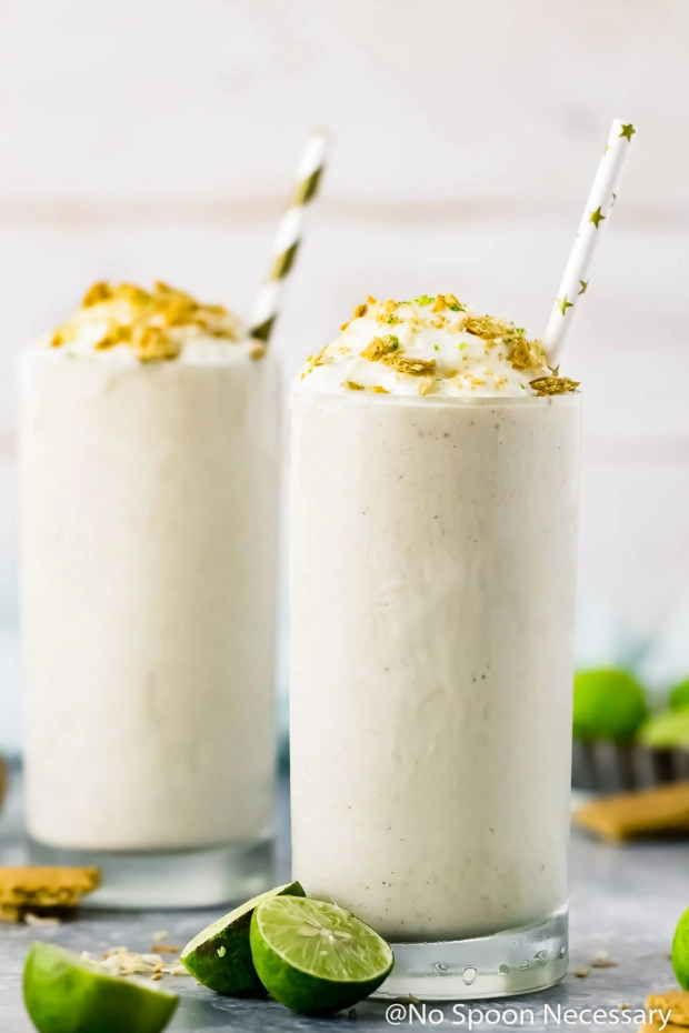 Straight on shot of two Lime Milkshakes topped with whipped cream, crushed graham crackers and lime zest with halved key limes surrounding the glasses.