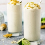 Straight on shot of two Lime Milkshakes topped with whipped cream, crushed graham crackers and lime zest with halved key limes surrounding the glasses and a bottle of tequila in the background.