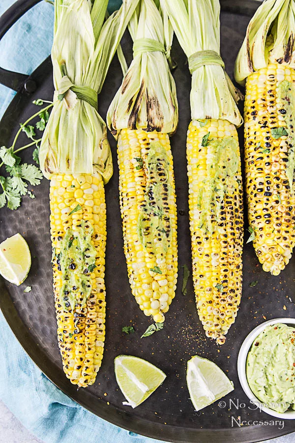 Overhead, up close shot of Grilled Corn with Avocado Sriracha Butter on a black platter with lime wedges and fresh cilantro.