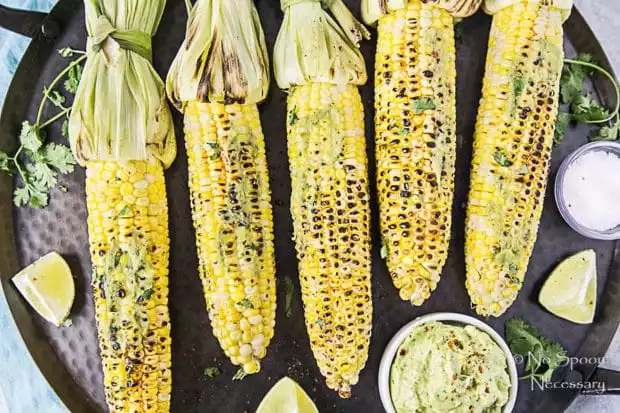 Overhead shot of Grilled Corn with Avocado Sriracha Butter on a black platter with lime wedges, fresh cilantro and a small bowl of salt.