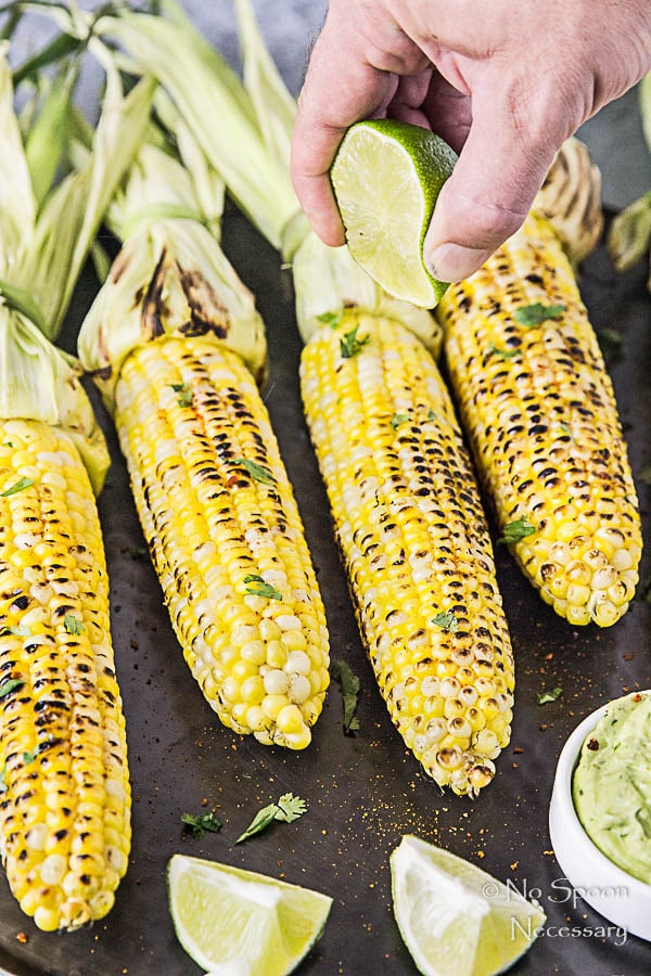 Angled shot of a hand holding a lime wedge and squeezing is over Grilled Corn with Avocado Sriracha Butter on a black platter. 