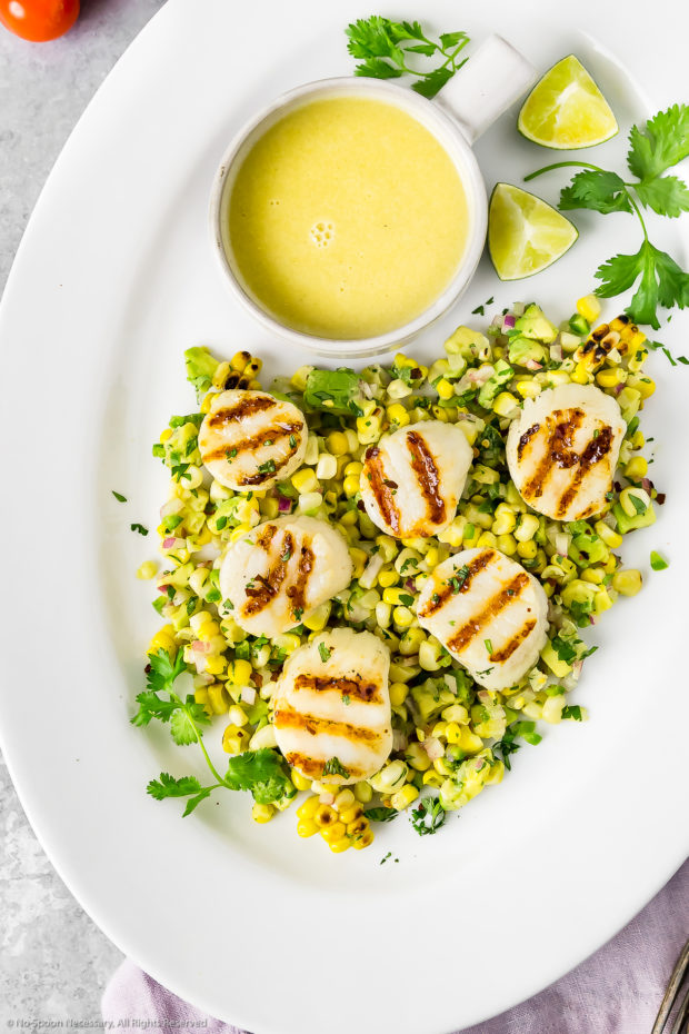 Overhead photo of Grilled Scallops served on a bed of Avocado & Corn Salsa on a white platter with a small bowl of Honey Lime Vinaigrette, fresh cilantro and lime wedges on the corner of the platter.
