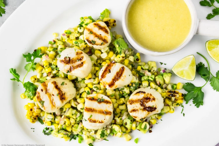 Overhead, landscape photo of Grilled Scallops on a bed of Avocado & Corn Salsa on a white platter with a small bowl of Honey-Lime Vinaigrette, fresh cilantro and lime wedges.