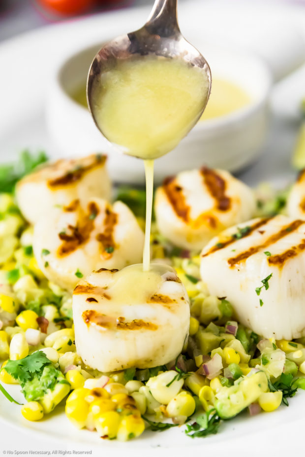 Straight on photo of a spoon drizzling honey lime sauce overtop a Grilled Scallop on a bed of Avocado and Corn Salsa.