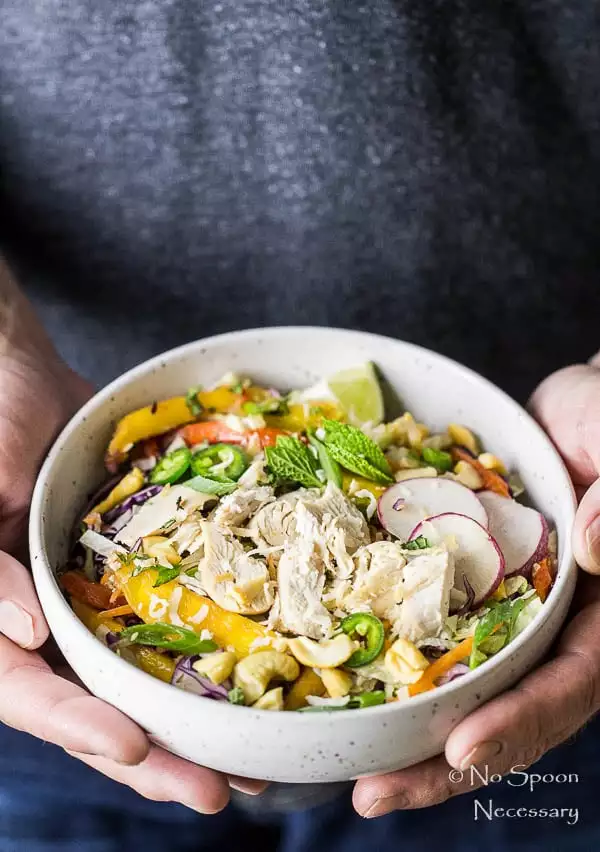 Angled photo of two hands holding a bowl of thai salad with mangoes and topped with shredded chicken.