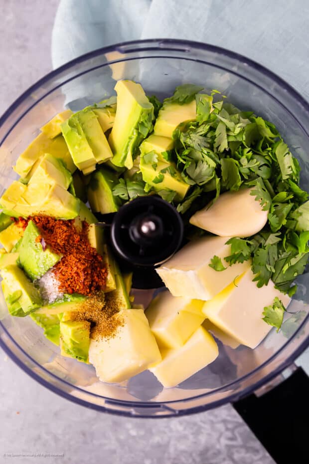 Overhead photo of chopped avocados, cubed unsalted butter, chopped cilantro, and dry seasonings in the bowl of a food processor.