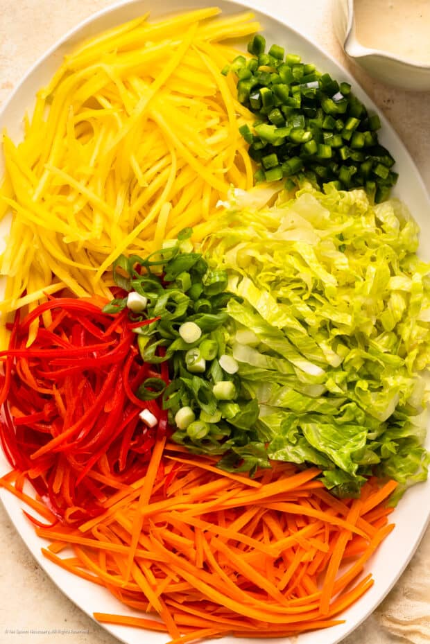 Overhead photo of julienned mango, shredded lettuce, strips of red bell pepper, julienned carrots, diced jalapenos and sliced scallions neatly organized on a white platter.