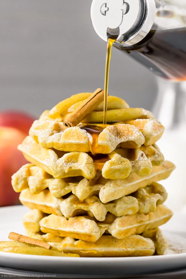 Straight on photo of a stack of Apple Cinnamon Waffles on a white plate with Maple Syrup in a glass jar being poured on top of the waffles.