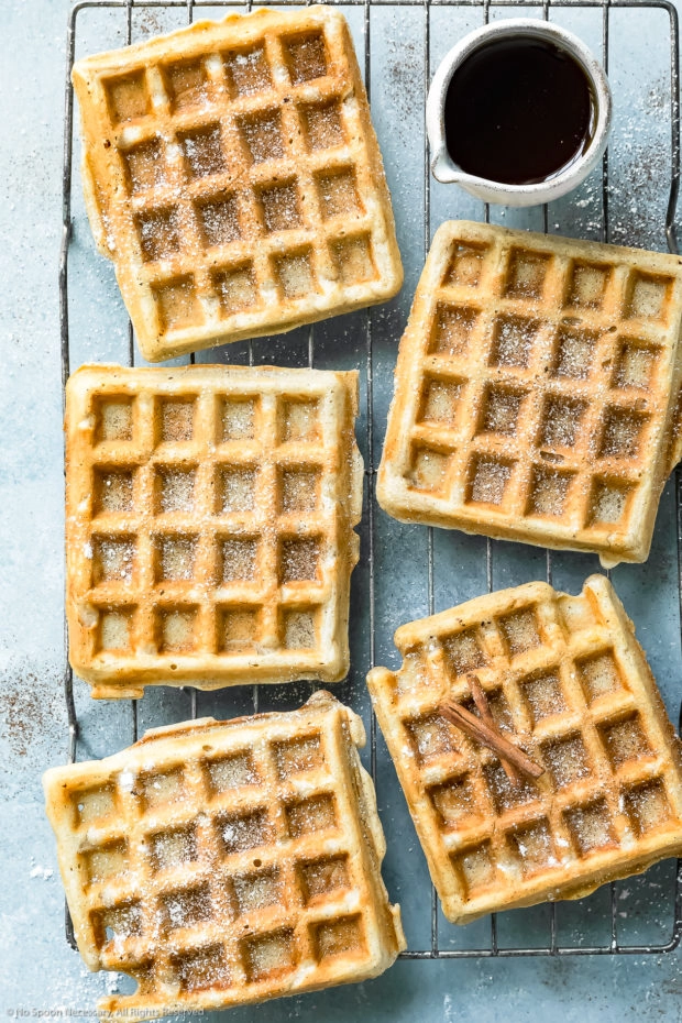 Overhead photo of Belgian cinnamon waffles dusted with powdered sugar and cinnamon on a wire cooling rack with a small ramekin of pure maple syrup off to the side.
