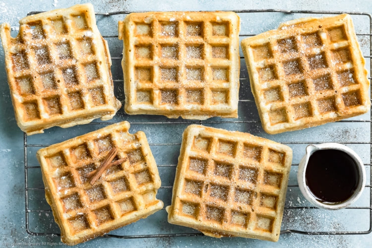 Overhead photo of Apple Cinnamon Waffles dusted with ground cinnamon and powdered sugar on a wire cooling rack with a ramekin of pure maple syrup.