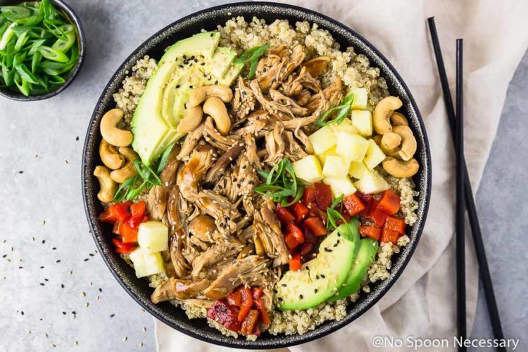 Overhead landscape shot of Slow Cooker Chicken Teriyaki Quinoa Bowls with black chop sticks on a silver board with a neutral linen and bowl of sliced scallions.
