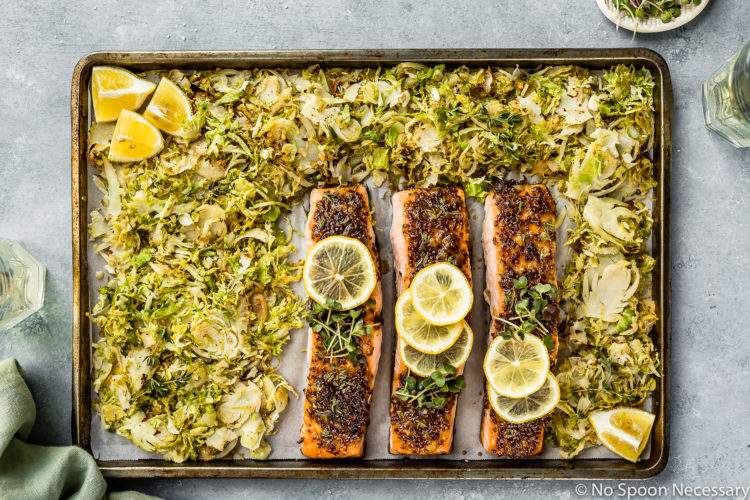 Overhead shot of Baked Honey Dijon Salmon with Brussels Sprouts on a sheet pan with a light green linen, glasses of wine and ramekin of microgreens surrounding the pan.