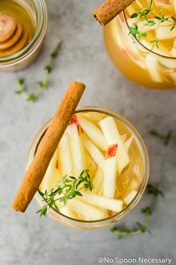 Overhead shot of a clear glass containing a Apple and Gin Autumn Cocktail garnished with matchstick cut apples, fresh thyme, and a cinnamon stick with more fresh thyme, a clear glass of honey and an additional cocktail surrounding.